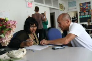 person assisting another writing in a notebook at a desk